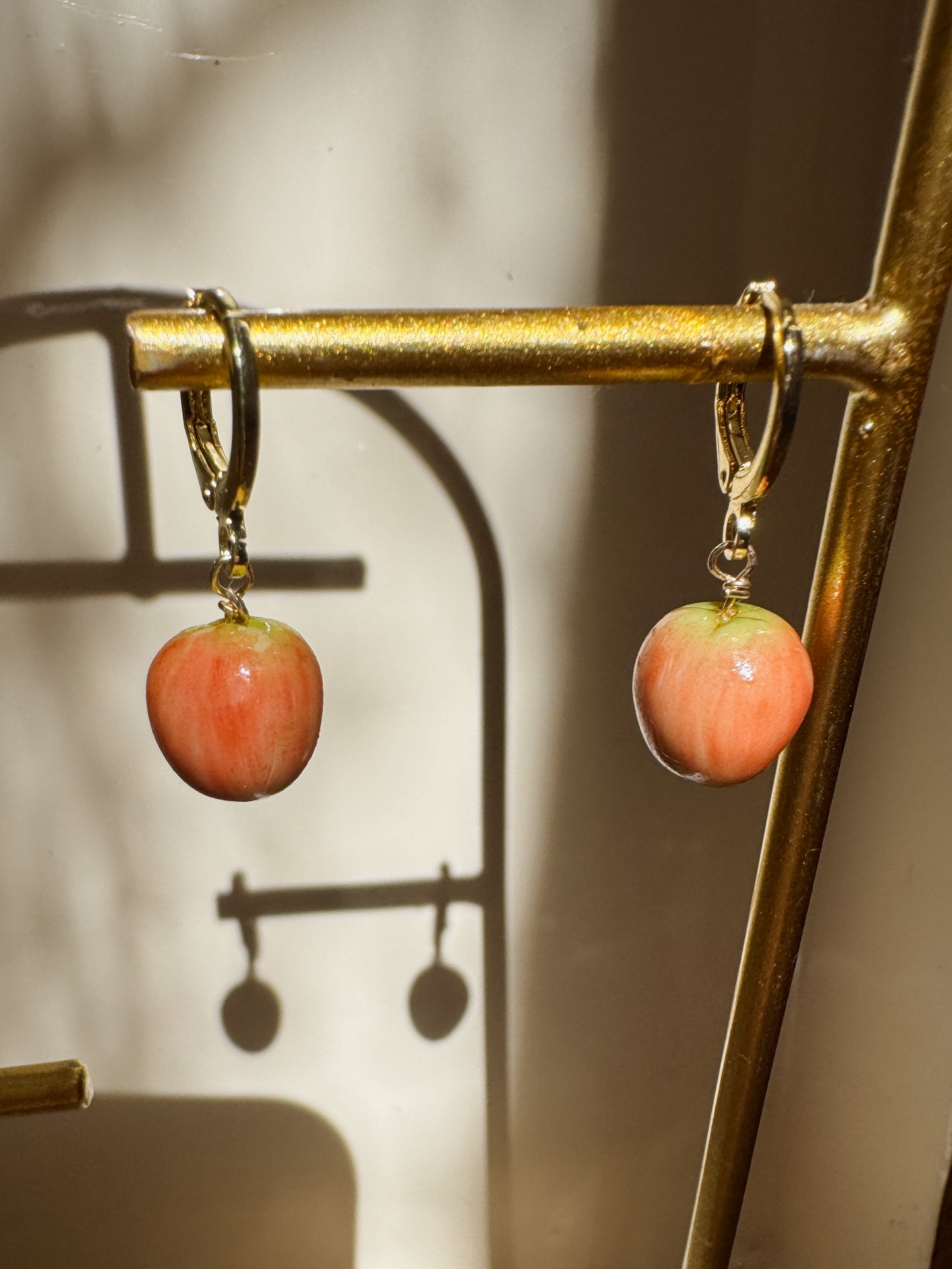 A pair of gold hoop earrings with dangling peach-colored apple charms, hanging from a gold display stand in warm, direct sunlight with strong shadows.