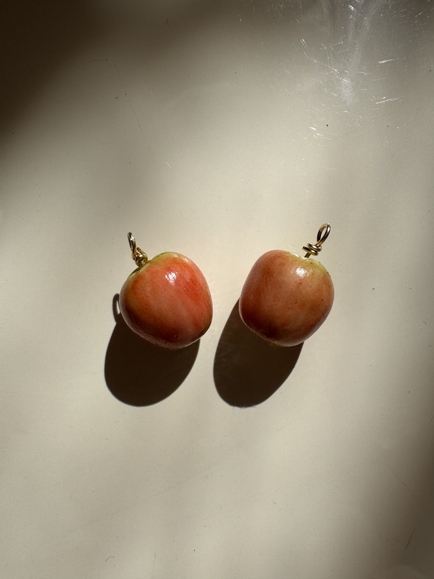 A pair of apple-shaped earrings with glossy reddish-brown resin bodies and small gold-tone hooks, photographed on a plain light surface with strong directional shadows.