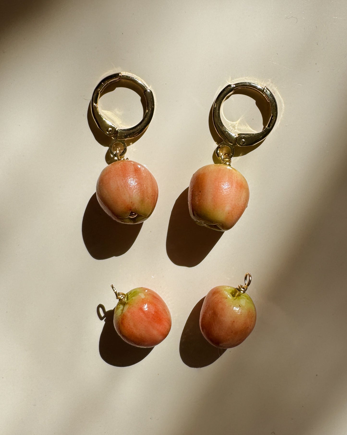 Three pairs of gold hoop earrings with small peach-pink apple charms, arranged on a warm beige surface with dramatic diagonal shadows.