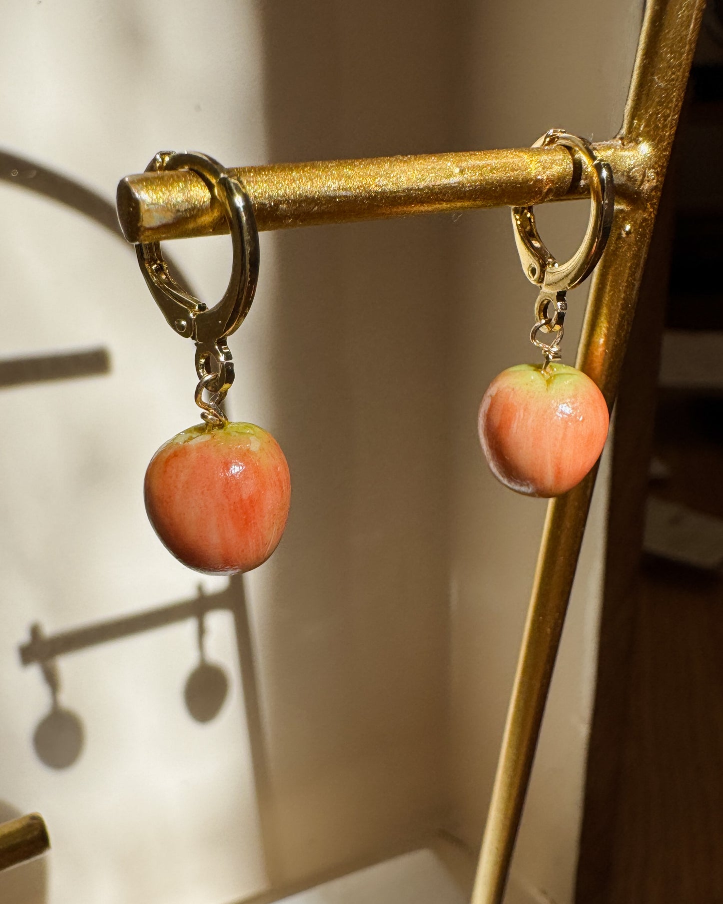 A pair of dangling earrings with peach-pink apple-shaped charms and gold-tone hoops, hanging from a gold display stand.