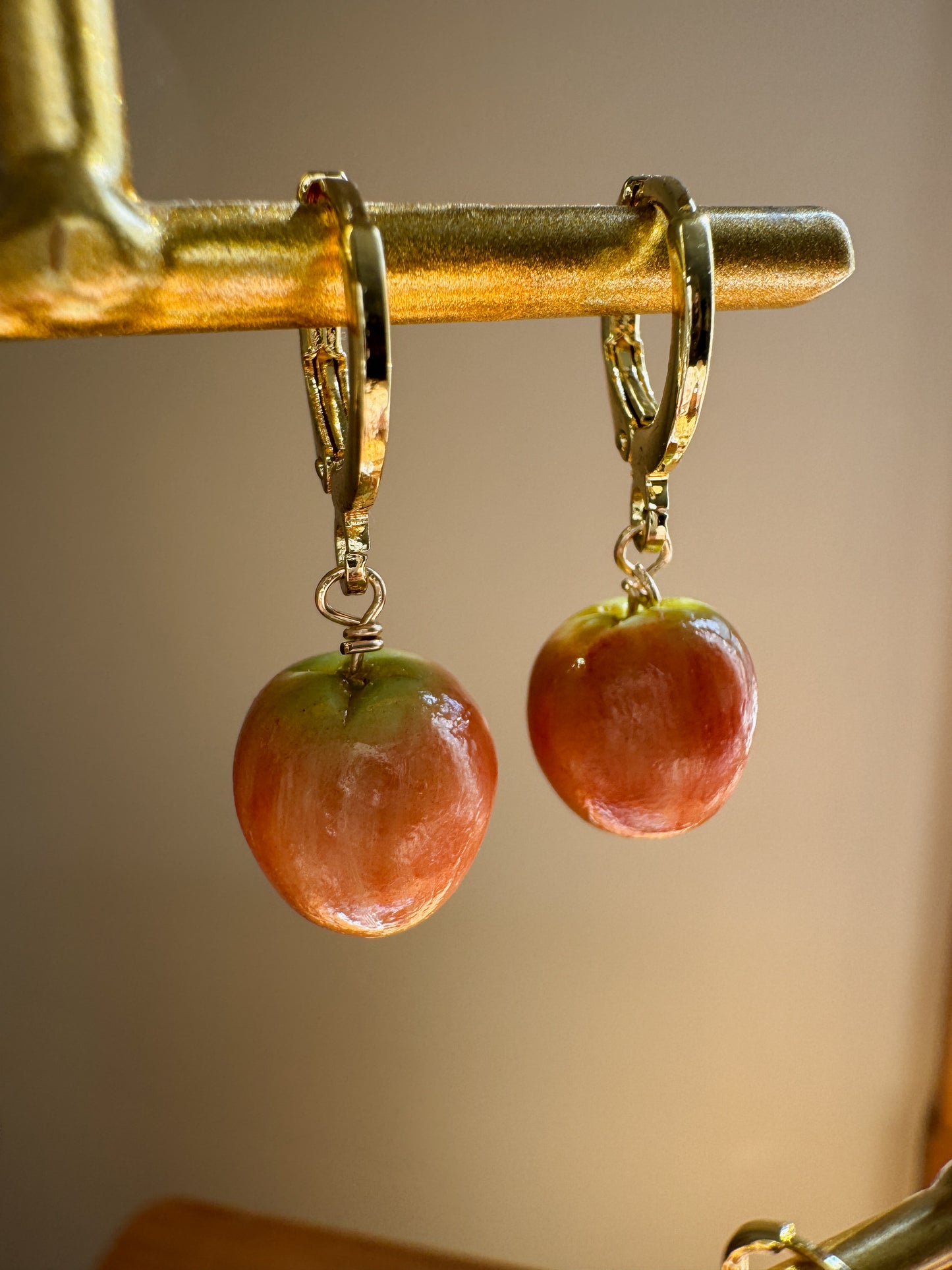 A pair of dangling earrings shaped like glossy red apples with green tops, hanging from gold-tone hoops on a gold display stand against a warm beige background.