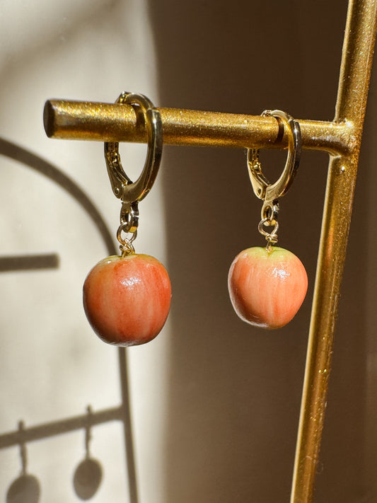 A pair of gold hoop earrings with dangling apple charms, displayed on a gold stand against a warm beige background with soft shadows.
