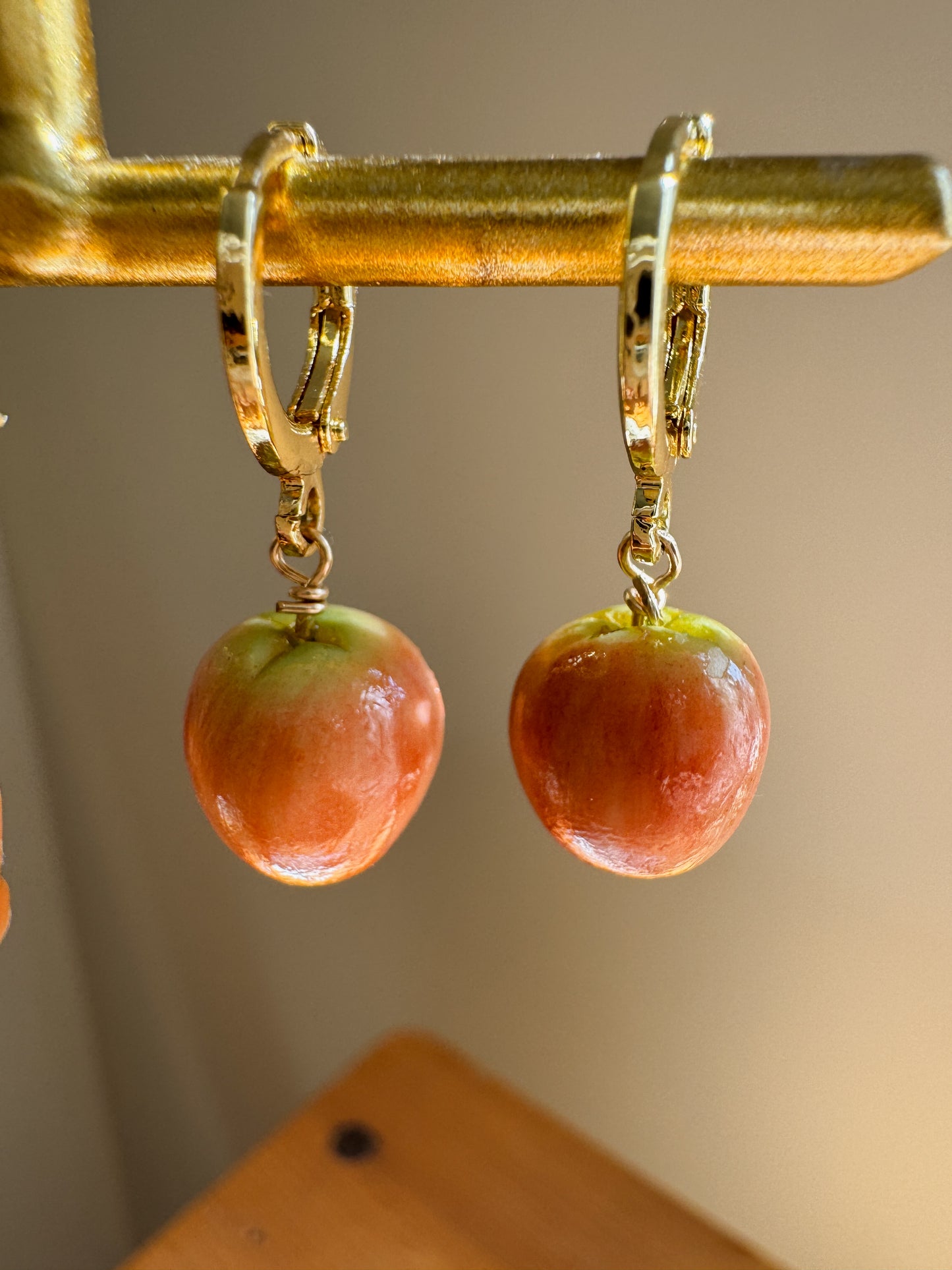 A pair of gold hoop earrings with dangling apple charms, hanging from a gold display stand against a warm beige background.